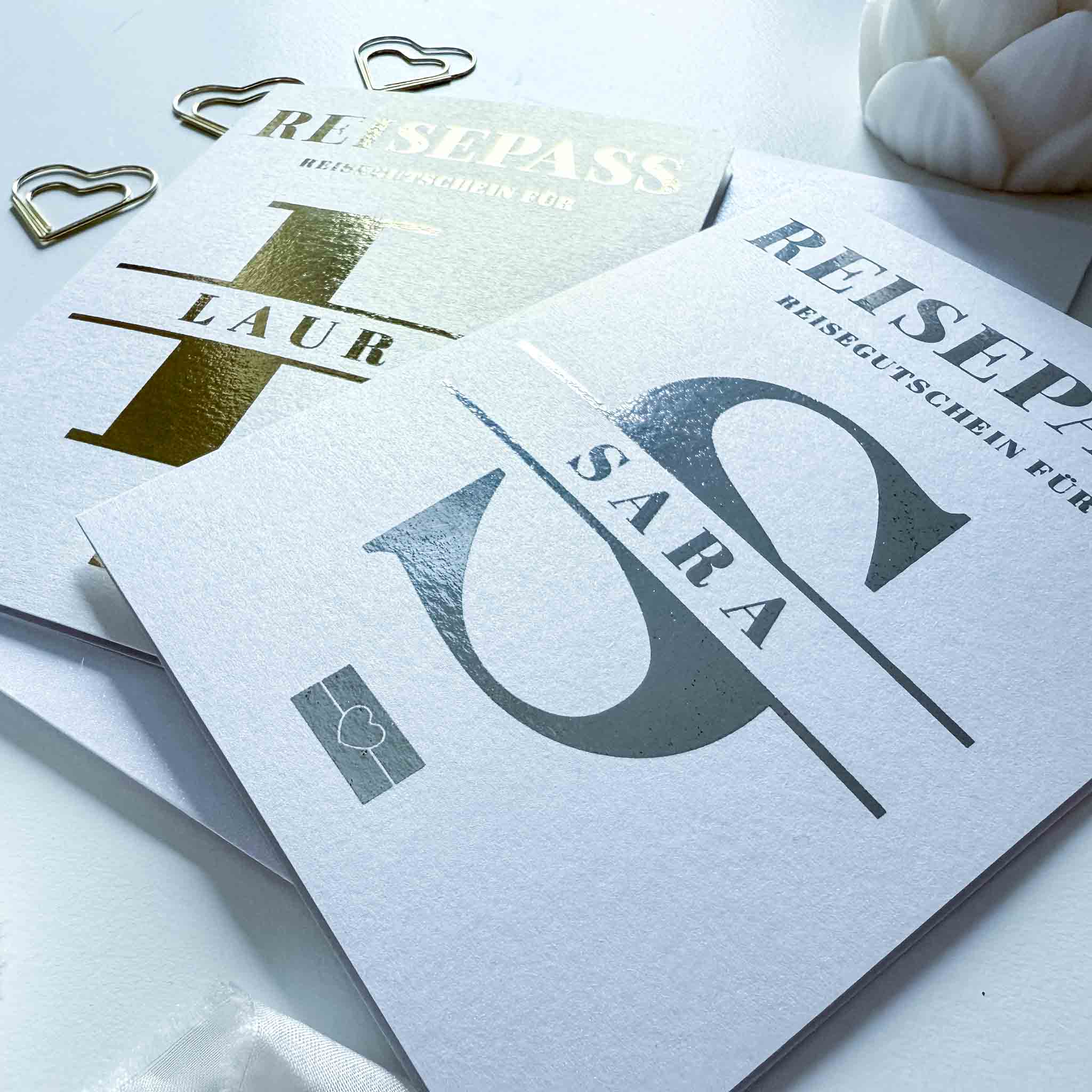 Zwei weiße funkenschauer Reisepass-Rubbelkarten als personalisierter Reisegutschein, eine mit "LAUR" in Gold, eine mit "SARA" in Silber. Mit Umschlag, goldener Herzklammer & Feder-die perfekte Geschenkidee für den Urlaub!.