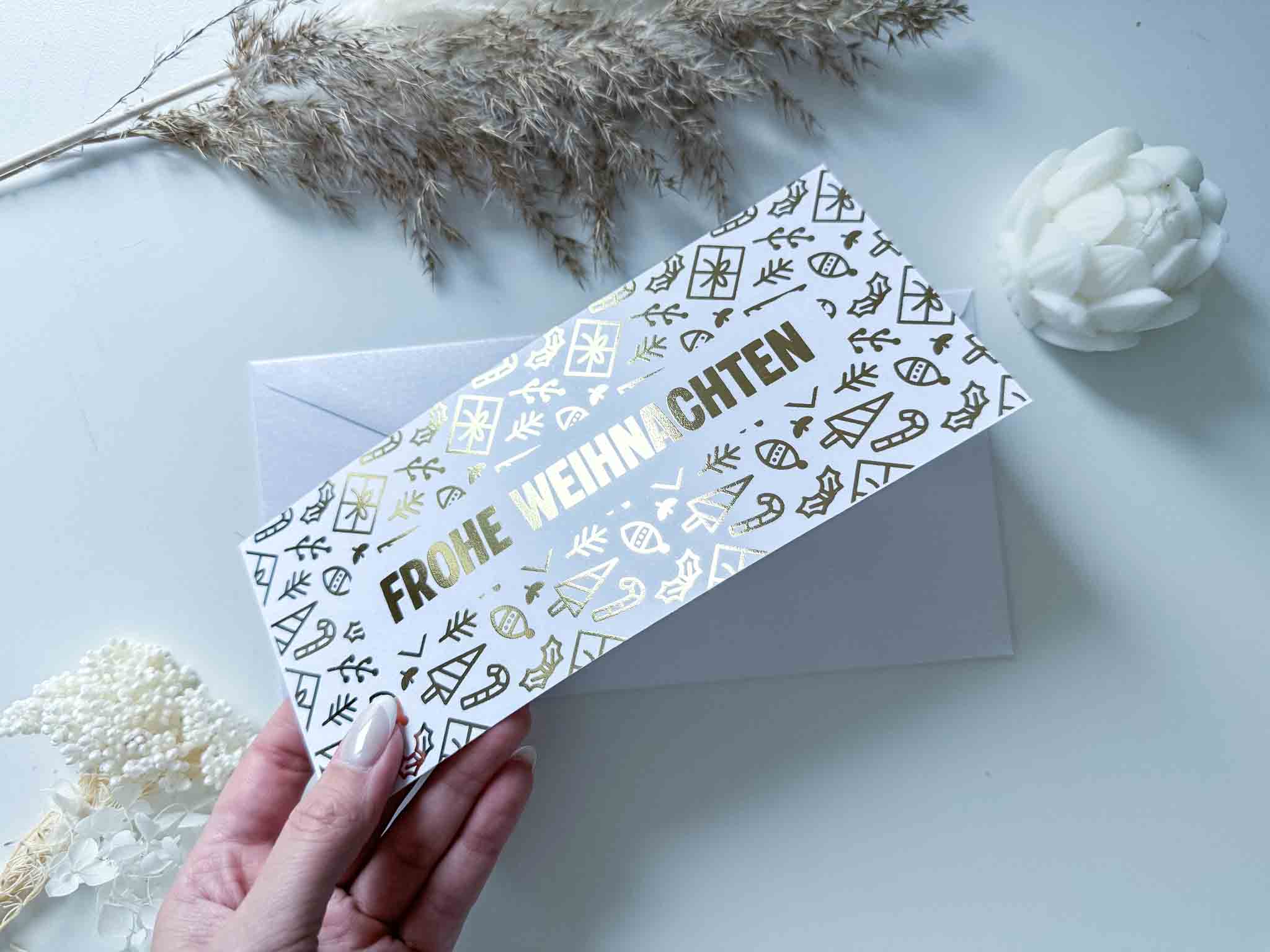 Eine Hand hält die funkenschauer Rubbelkarte "Karte Weihnachten Gutschein Reise personalisiert" mit goldenen Details und "Frohe Weihnachten", über einem weißen Umschlag, umgeben von getrockneten Pflanzen und einer dekorativen weißen Blume.