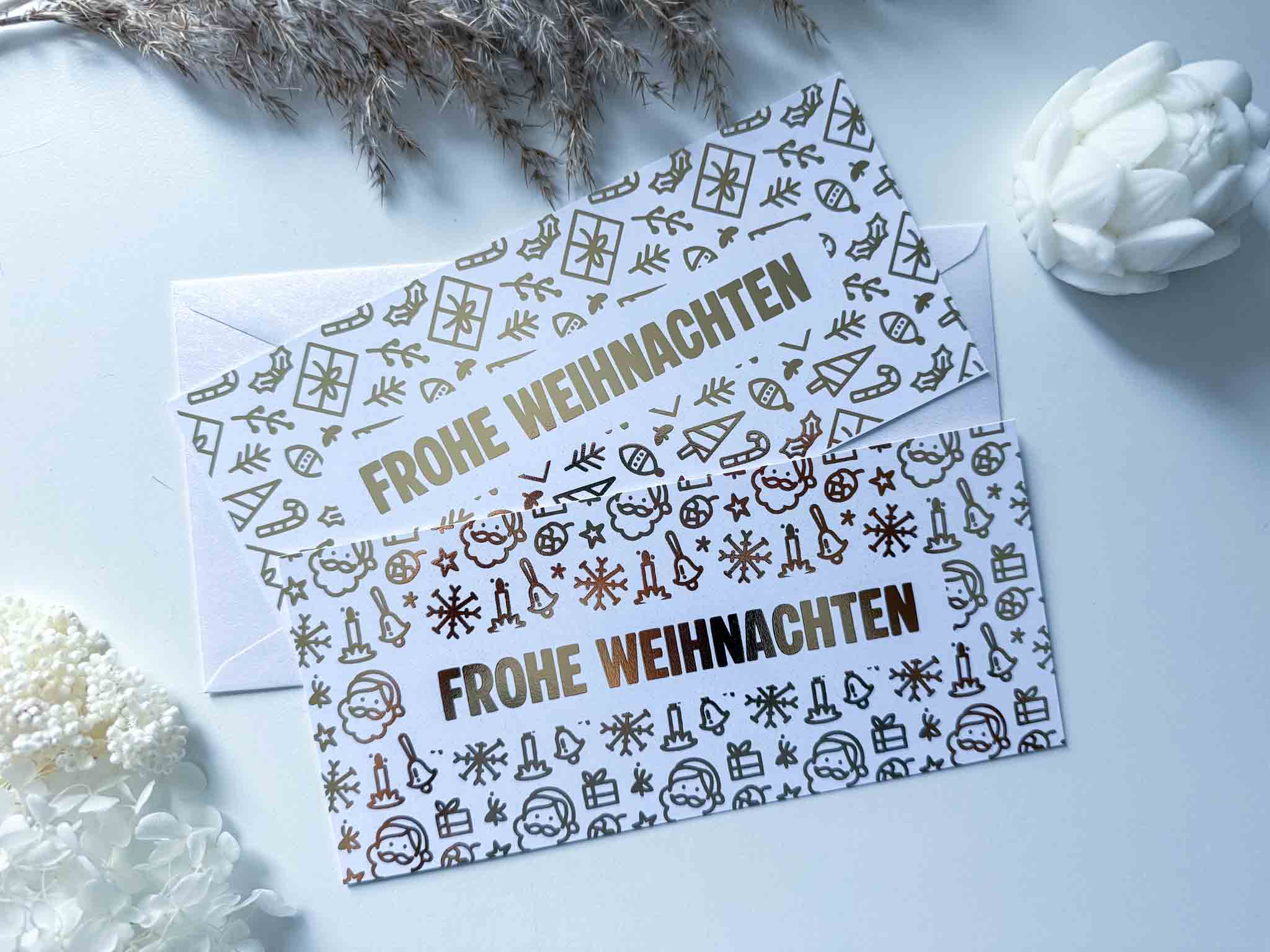 Zwei festlich gestaltete funkenschauer Rubbelkarten "Karte Weihnachten Gutschein Reise personalisiert" mit "Frohe Weihnachten", goldenen und schwarzen Symbolen, liegen auf einer weißen Fläche neben einem Briefumschlag, einer weißen Blume und getrockneten Pflanzen.