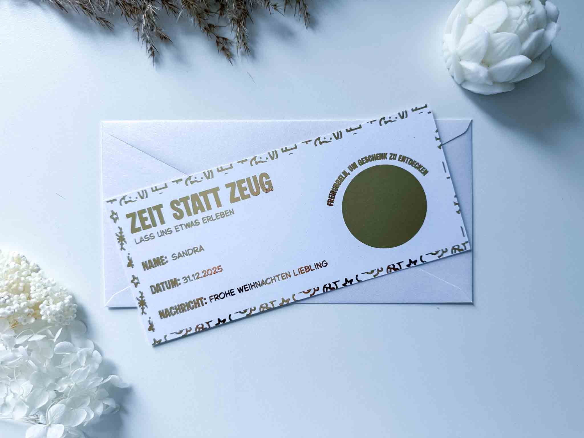 Die personalisierte Weihnachtsgutschein-Rubbelkarte "Zeit statt Zeug" von funkenschauer liegt auf einem weißen Umschlag, umgeben von weißen Blumen und Dekoration auf hellem Untergrund.