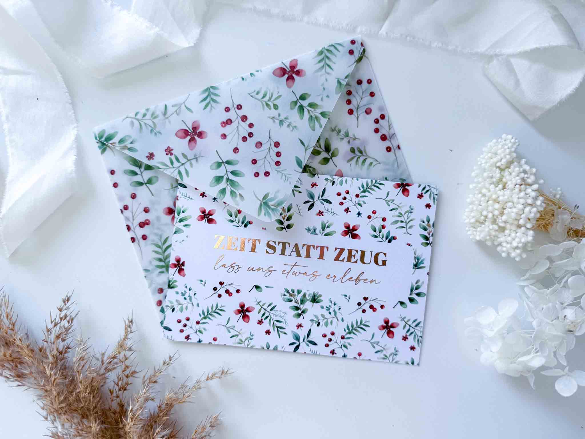 Die funkenschauer Weihnachten Gutschein "Zeit statt Zeug" Karte mit handgemachtem Umschlag liegt auf weißem Stoff, umgeben von Trockenblumen und weißen Beeren.