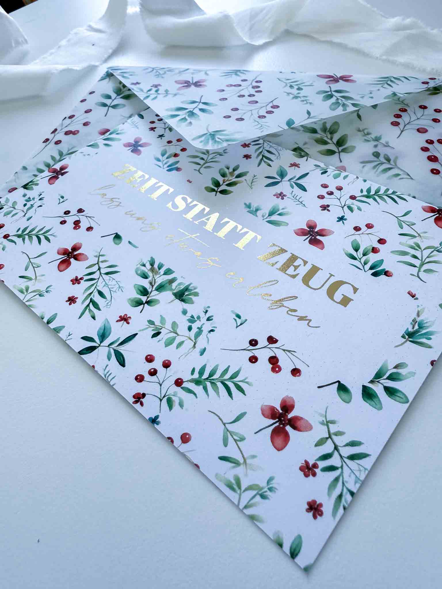 Ein handgefertigter floral gemusterter "Weihnachten Gutschein Zeit statt Zeug"-Umschlag von funkenschauer mit goldenem Text und Schrift liegt teilweise geöffnet auf einer weißen Fläche mit einem weißen Band im Hintergrund.