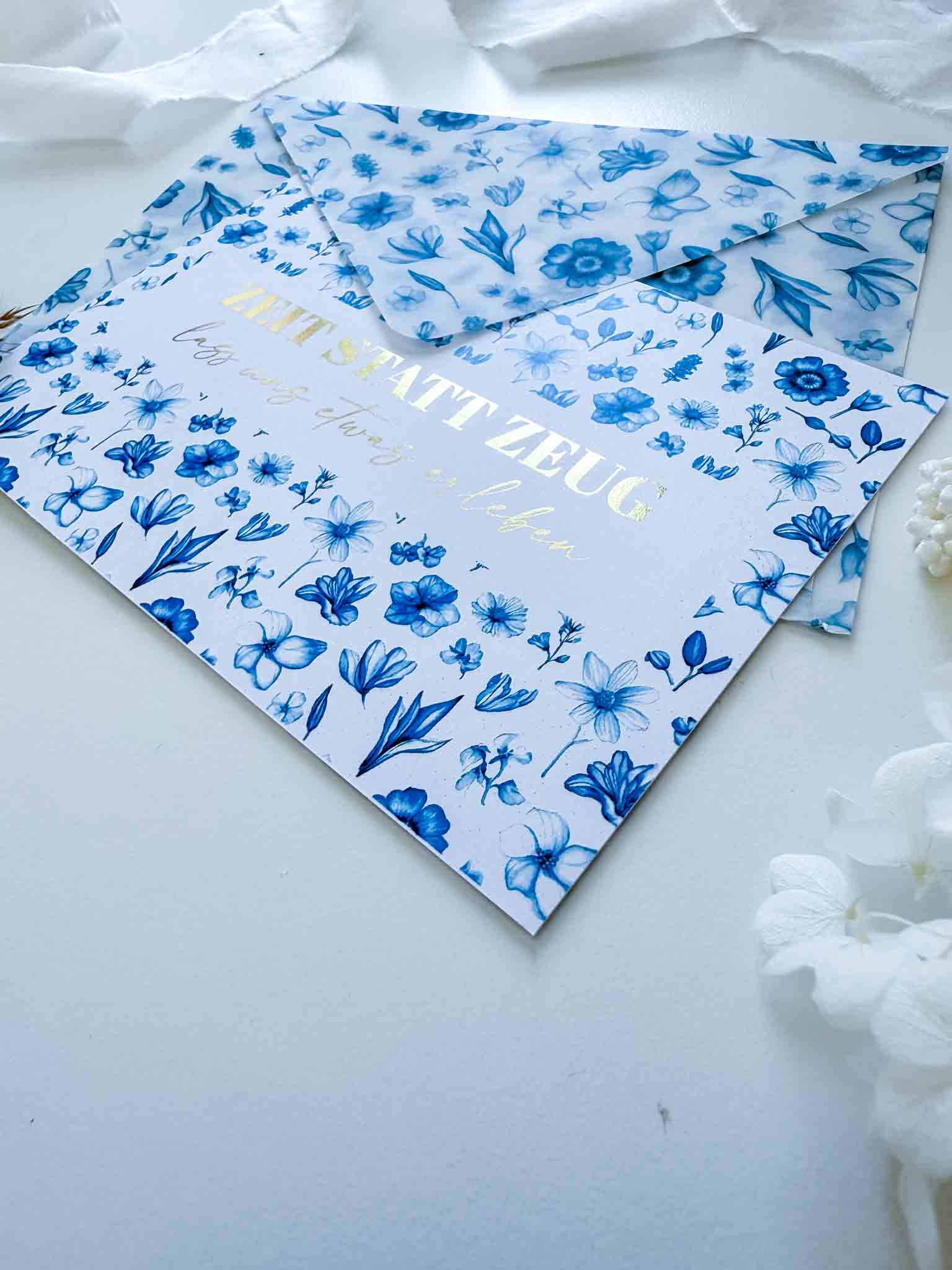 Ein Gutschein Zeit statt Zeug Rubbelkarte von funkenschauer, mit floralen Mustern und Goldfolie, steckt teils im handgemachten passenden Umschlag. Im Hintergrund sind weiße Blumen und Stoff auf einer weißen Oberfläche zu sehen.