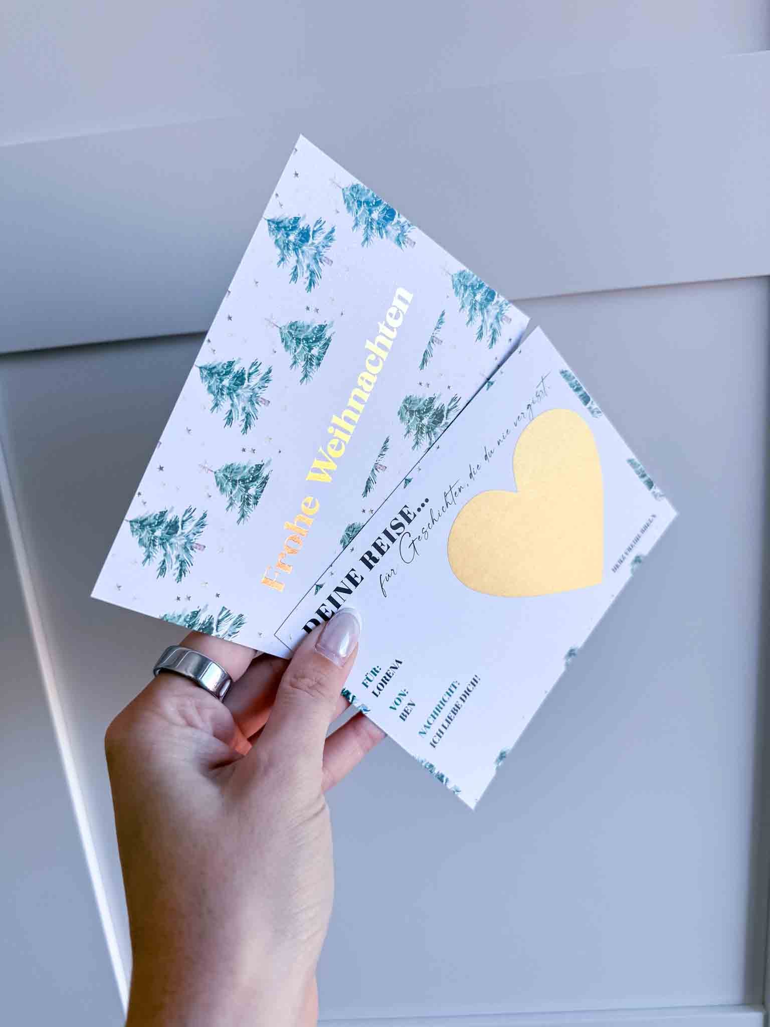 Eine Hand hält die funkenschauer "Reisegutschein Weihnachten"-Rubbelkarte mit grünen Bäumen, goldenem "Frohe Weihnachten"-Schriftzug und einem goldenen Herz - perfekt als personalisiertes Weihnachtsgeschenk - vor einem hellgrauen Hintergrund.