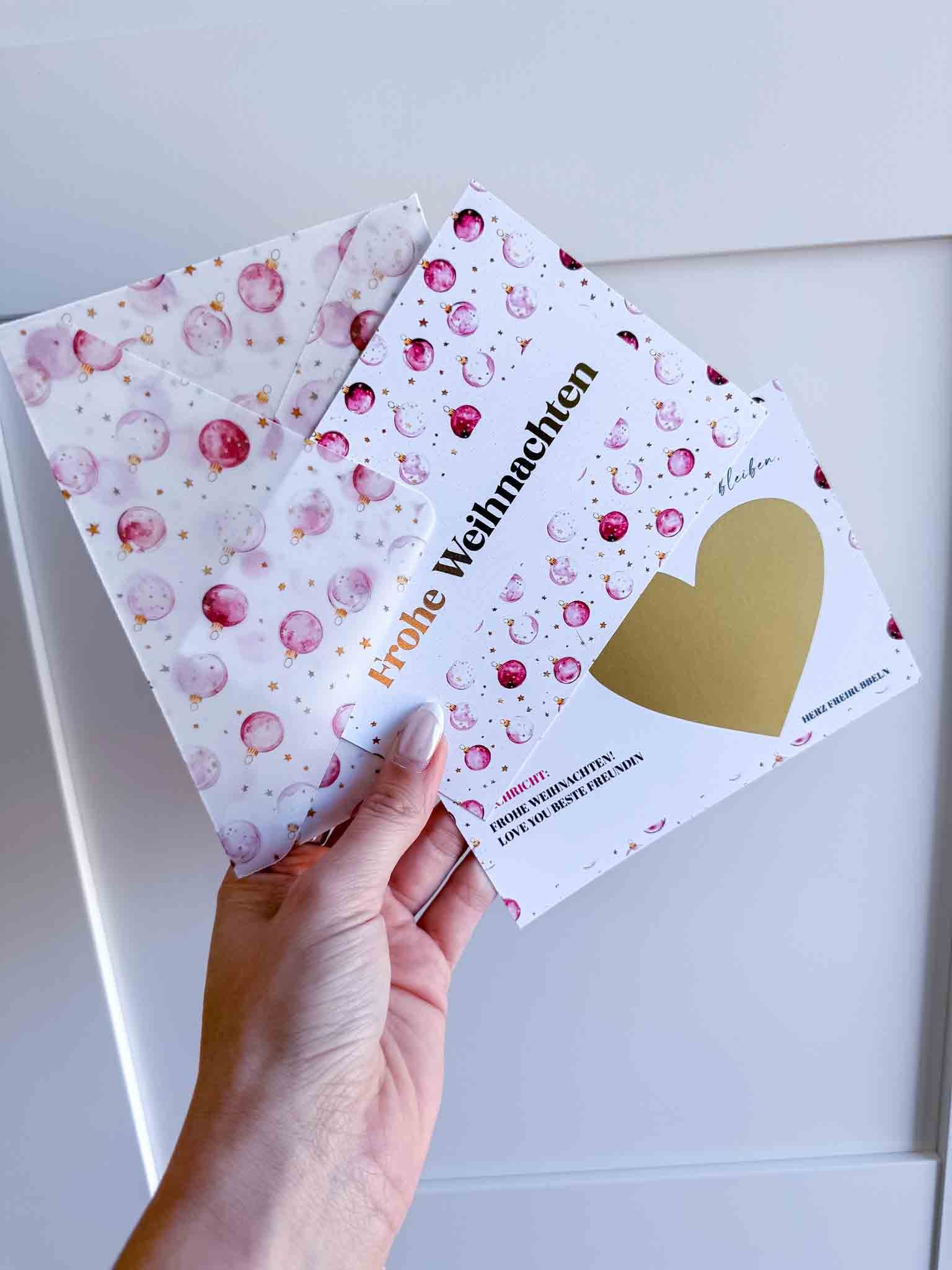 Eine Hand hält einen funkenschauer Weihnachten Gutschein Zeit statt Zeug Rubbelkarte mit "Frohe Weihnachten", pinken Ornamenten und goldenen Sternen - inklusive handgemachtem Umschlag, ideal als Weihnachtsgeschenk oder Gutschein.