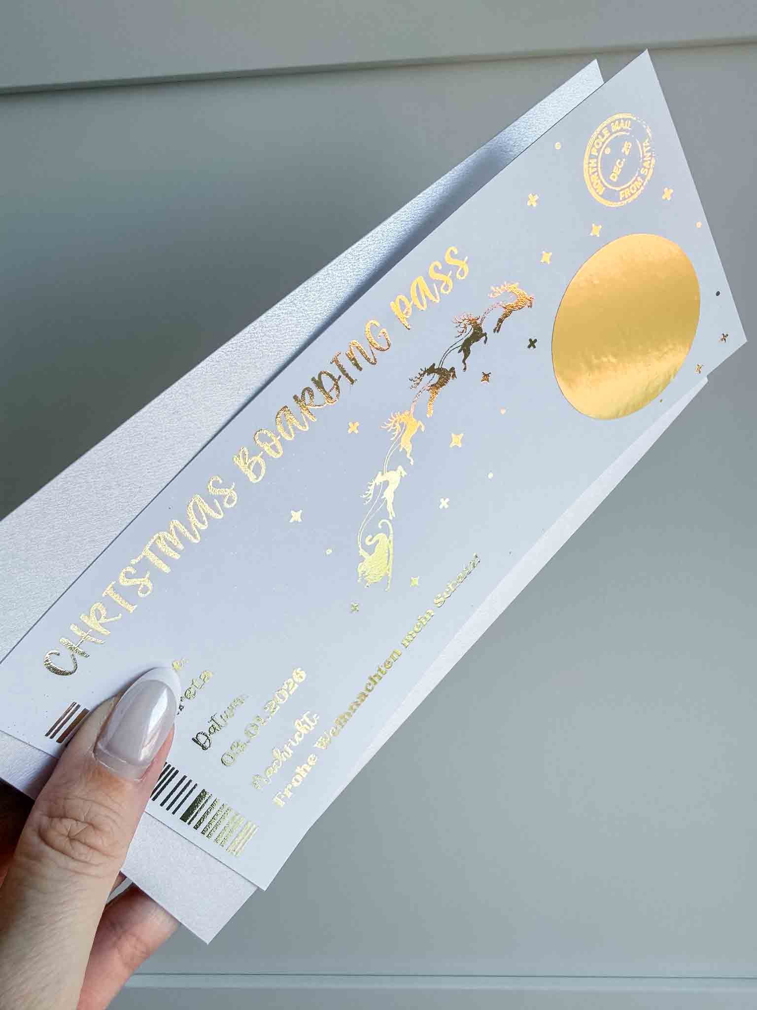 Eine Hand mit hellem Nagellack hält den funkenschauer Weihnachtsgutschein Reisegutschein Boarding Pass - eine personalisierte Rubbelkarte mit goldener Folie, Rentierschlitten und Sternen. Perfektes Geschenk Reise oder Flugticket zu Weihnachten!.