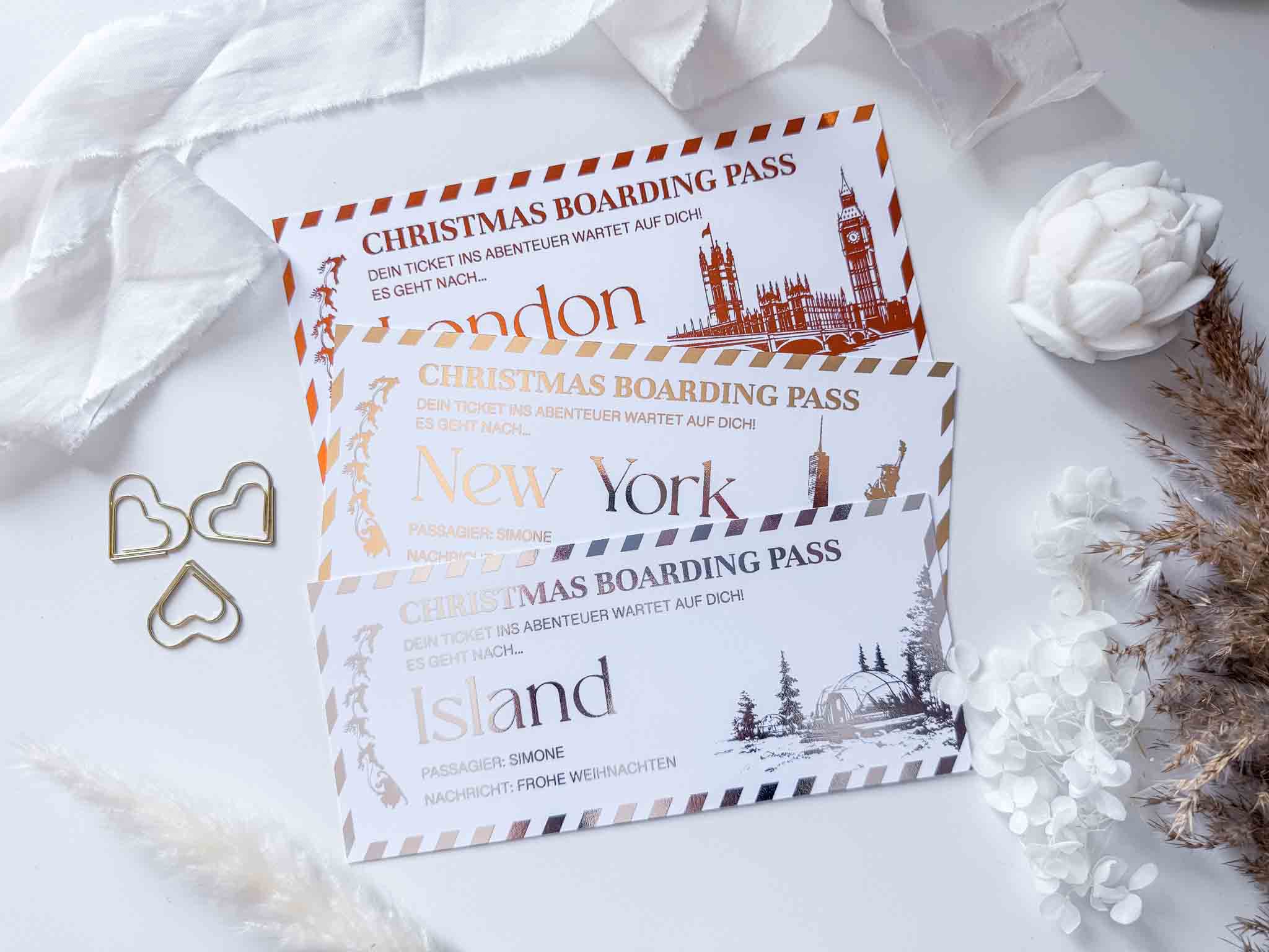 Drei "Weihnachtsgutschein"-Karten von funkenschauer, personalisiert für London, New York & Island, liegen auf weißem Untergrund mit Trockenblumen und goldenen Clips-perfekt als origineller Reisegutschein als Weihnachtsgeschenk.