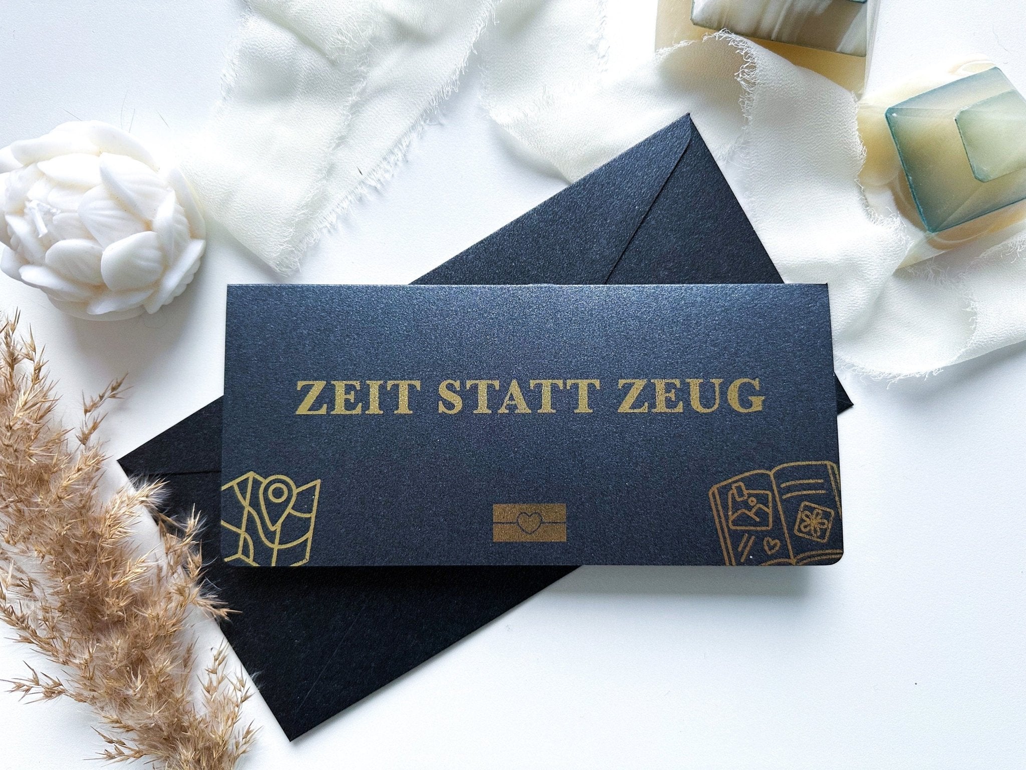 Ein „Gutschein Zeit statt Zeug personalisiert mit Rubbelfeld | Geschenk Aktivität, gemeinsame Zeit | Weihnachten“ von Funkenschauer, ausgestattet mit einem schwarzen Sonderedition-Umschlag mit goldenem Schriftzug „ZEIT STATT ZEUG“, wird auf eine weiße Fläche gelegt, die mit Dekorationsgegenständen wie einer weißen Blume verziert ist , beiger Stoff und getrocknetes Pampasgras. Dieser elegant gestaltete Umschlag zeigt goldene Symbole eines Globus, eines Herzens in einer Schachtel und eines Buches mit Herz.