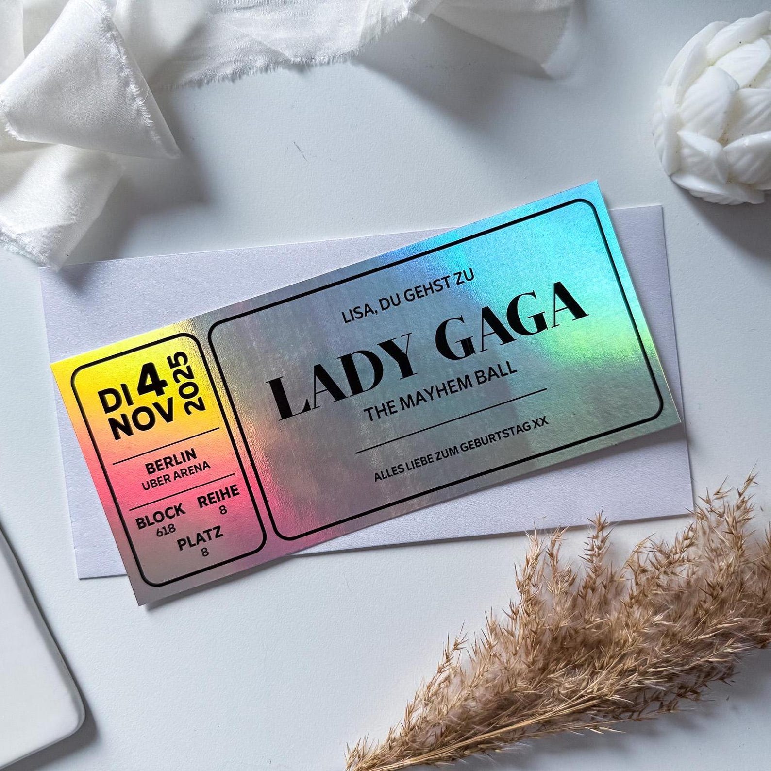 Ein funkenschauer Konzert Gutschein personalisiert für Lady Gaga's "The Mayhem Ball" am 4.11.2025 in Berlin liegt auf einem weißen Umschlag-ein perfektes Konzert Geschenk-umgegeben von weißem Stoff, Trockenblumen und einem weißen Keyboard.