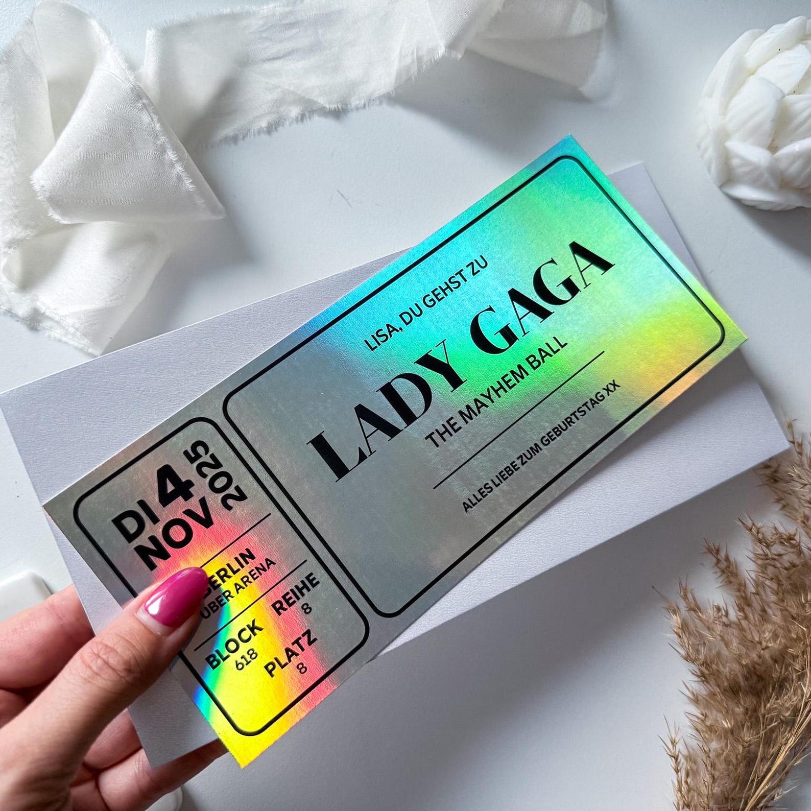 Eine Hand hält einen funkenschauer Konzert Gutschein personalisiert für Lady Gagas "The Mayhem Ball" in Berlin (4. November 2025), gedruckt auf holografischem Papier, mit weißem Stoff und Blumen im Hintergrund.