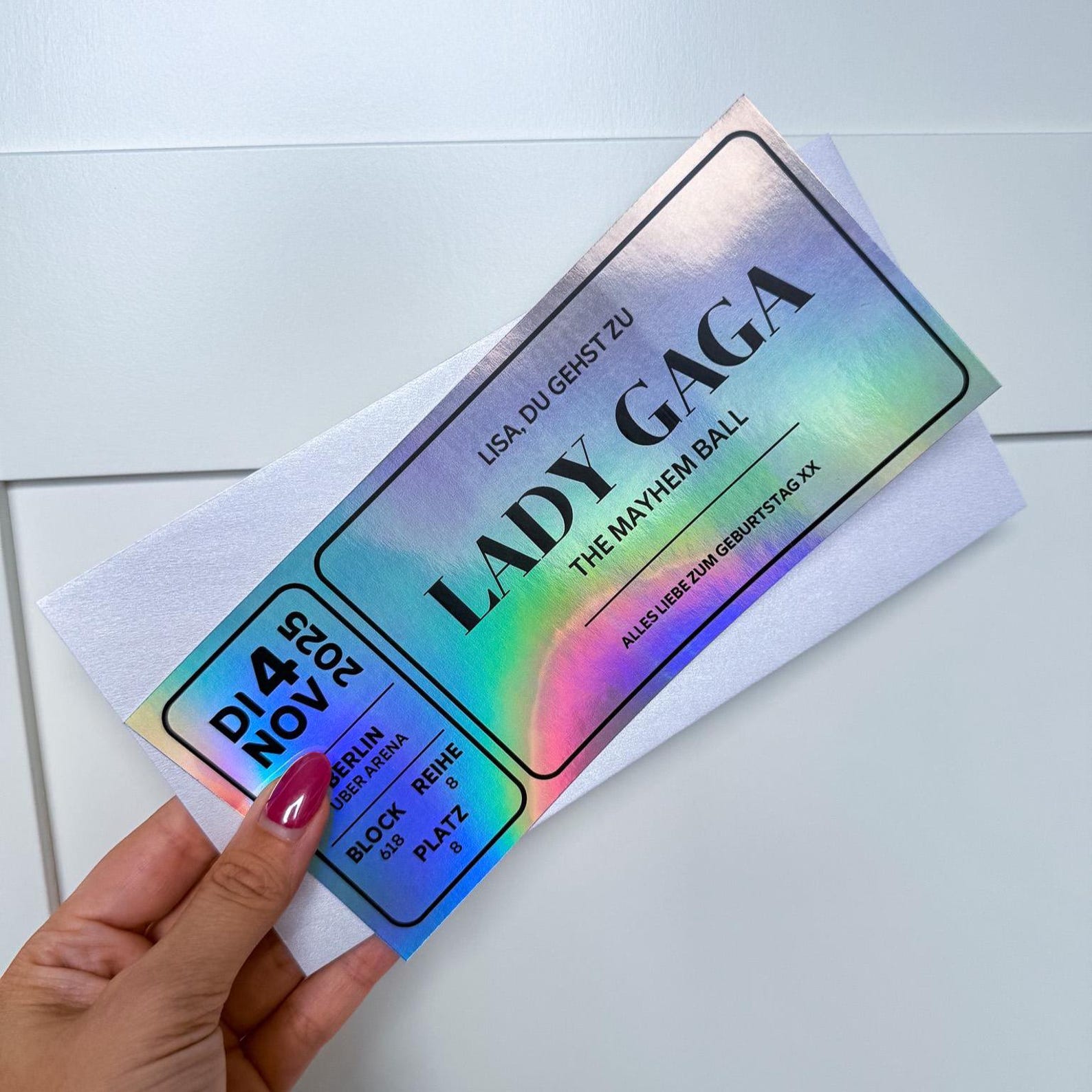 Eine Hand mit rosafarbenen Nägeln hält einen funkenschauer Konzert Gutschein personalisiert - einen personalisierten Konzertgutschein - in deutscher Sprache vor einem weißen Hintergrund.