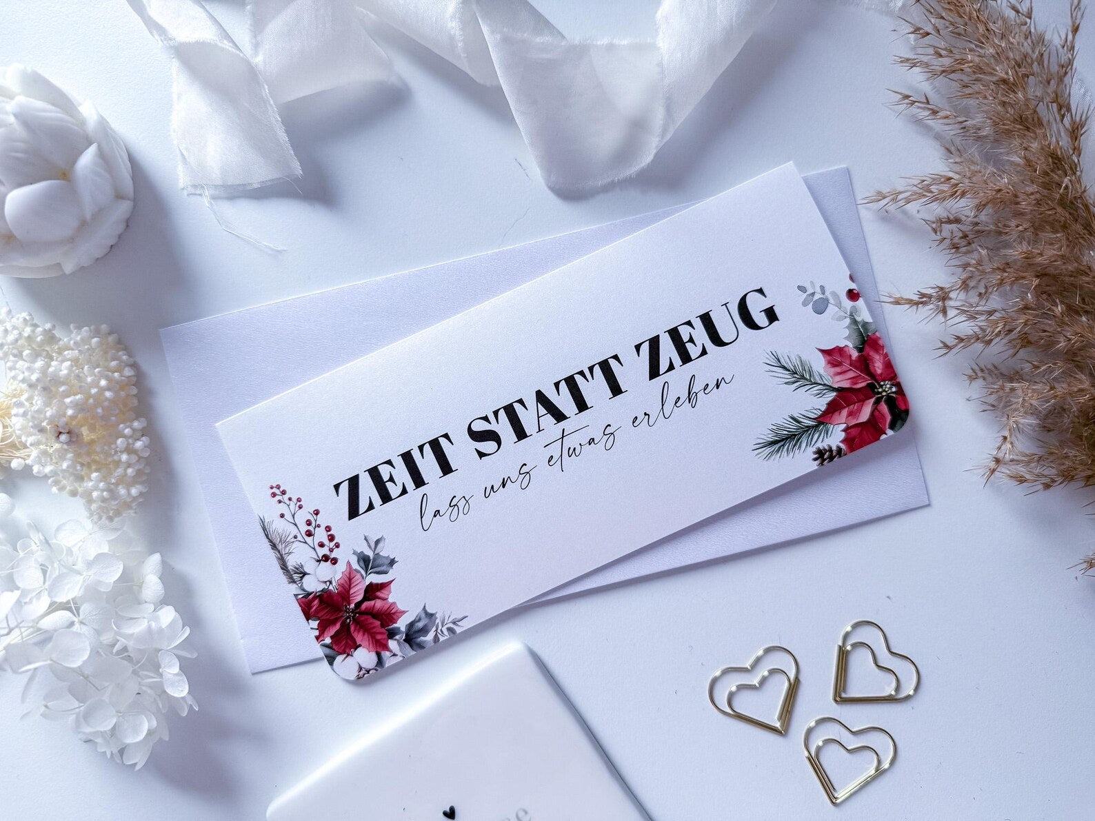 Eine weiße "Zeit statt Zeug Gutschein Weihnachten" Karte von funkenschauer mit Blumen und dem Spruch "ZEIT STATT ZEUG, lass uns etwas erleben" liegt auf einem Umschlag, umgeben von Deko - das perfekte personalisierte Weihnachtsgeschenk!.