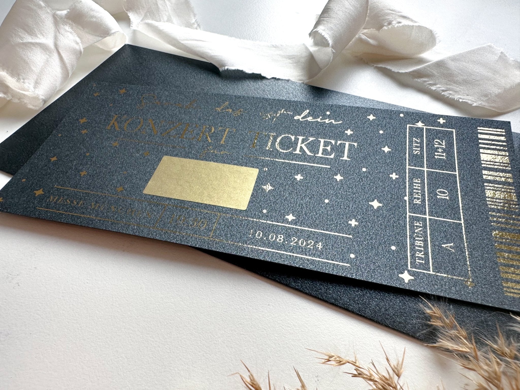 Ein personalisiertes Konzertticket von funkenschauer, schwarz mit goldener Schrift und mit Sternen- und Mondmotiven, liegt auf einer weißen Fläche neben weißen Stoffstücken und getrocknetem Gras. Das Ticket beschreibt ein Konzert am 10. August 2024 im Block A, Reihe 10, Platz H12 – eine Einladung zu unvergesslichen Musikmomenten für Fans von Ed Sheeran oder Wincent Weiss.