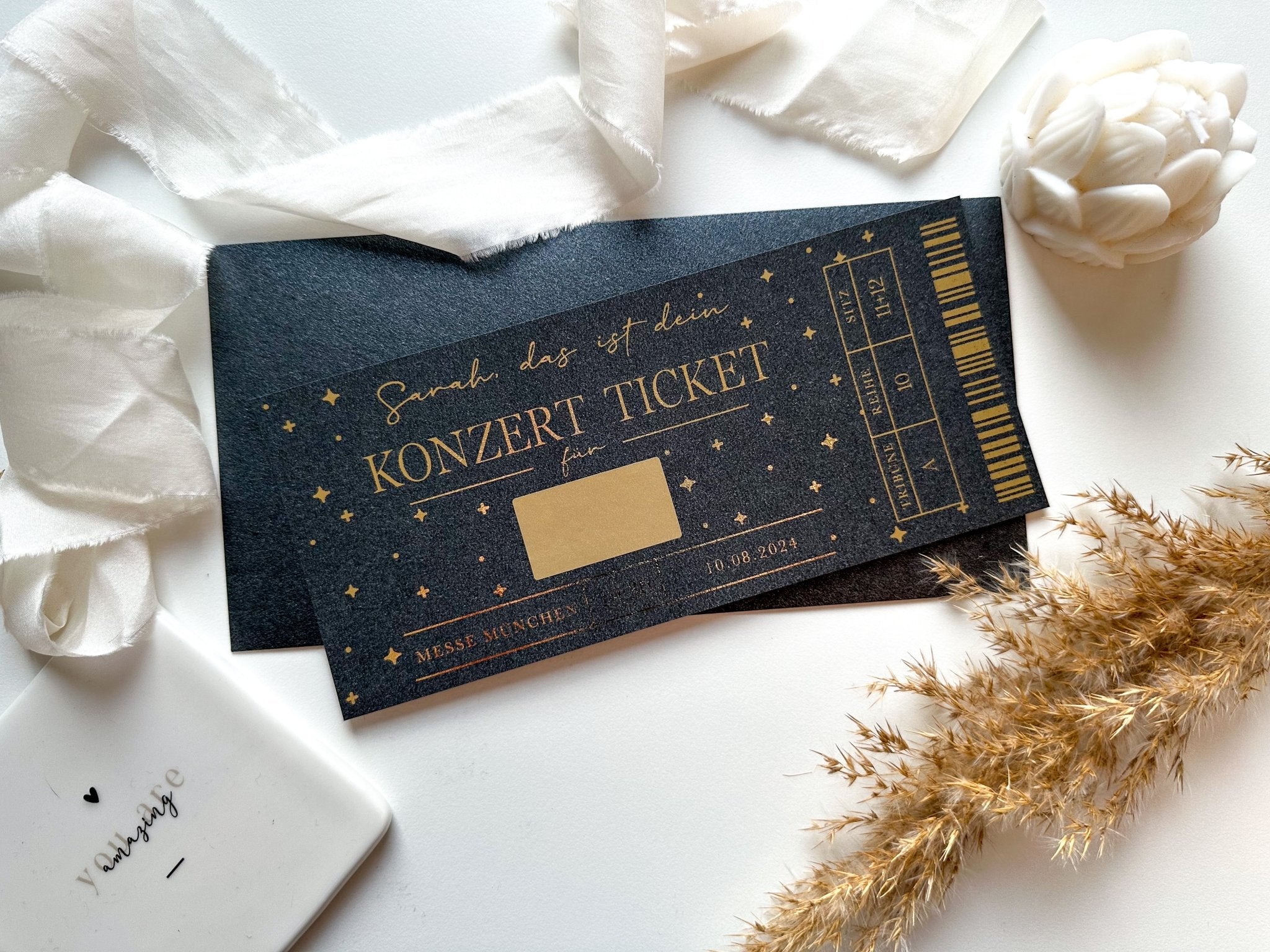 Ein personalisierbares Konzertticket von funkenschauer mit goldenem Text und Sternen auf schwarzem Hintergrund ist elegant von einem dekorativen weißen Band, einer weißen Blume, getrocknetem Gras und einer weißen Schale mit einem kleinen Herz umgeben. Auf dem Ticket steht „Schenk das ist dein KONZERTTICKET“ und die Angaben „MESSE MÜNCHEN“ sowie ein bestimmtes Datum – ein unvergessliches, personalisiertes Geschenk für jeden Musikliebhaber.