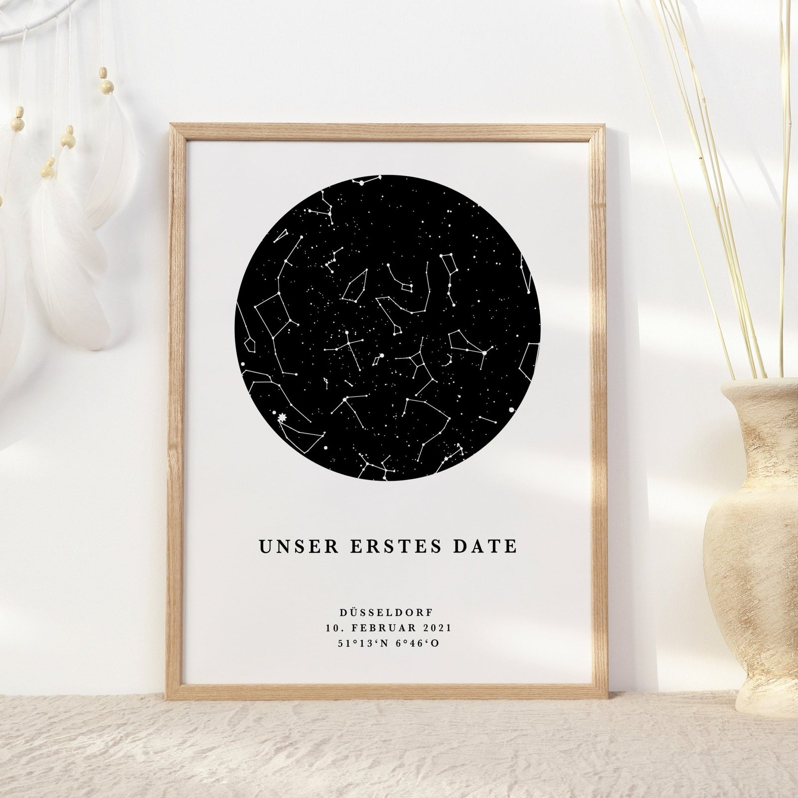 Ein gerahmtes Sternenhimmel-Poster personalisiert von Funkenschauer mit Sternbildern mit dem Titel „Unser erstes Date“ zeigt die Details: „Düsseldorf, 10. Februar 2021, 51°13'N 6°46'O.“ Es wird elegant auf einer hellen Oberfläche neben einer Vase und weißen Federn platziert.