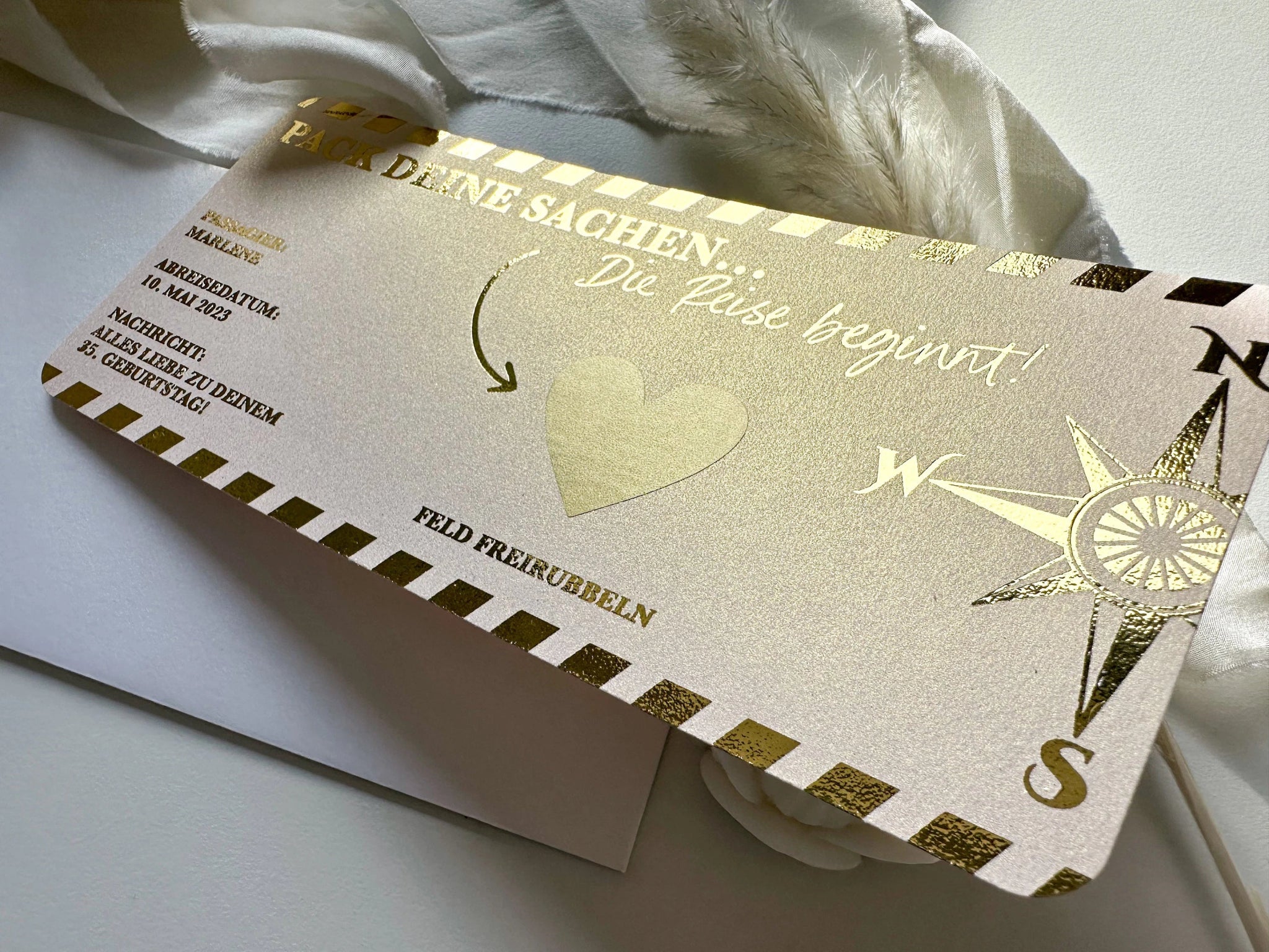 Ein personalisierter Reisegutschein mit Rubbelfeld, elegant präsentiert als Ticket-Einladung in Gold und Weiß mit Kompassdesign und gedruckt auf luxuriösem Roségold-Perlmuttpapier. Der Text in deutscher Sprache lautet: „Pack deine Sachen. Die Reise beginnt!“. Dieses exquisite Geschenk von funkenschauer enthält das personalisierte Geschenk, das teilweise auf einem weißen Umschlag ruht.