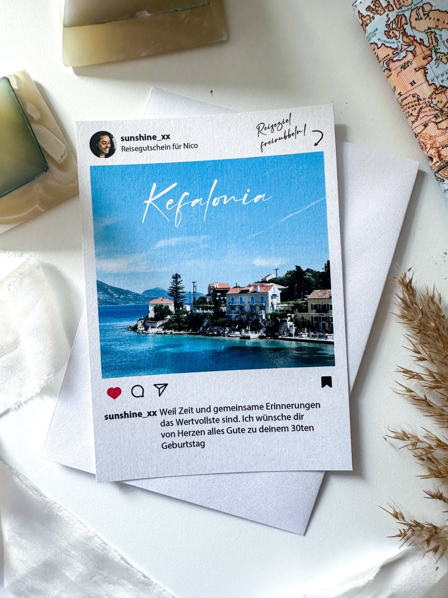 Eine Postkarte mit dem Bild einer Küstenstadt mit Häusern und Bäumen entlang der Küste, beschriftet mit „Kefalonia“. Die Postkarte liegt auf einem weißen Blatt Papier, neben einer Teilansicht einer Karte, einer Pflanze und anderen kleinen Dekorationsgegenständen. Der Text ist auf Deutsch. Perfekt als Reisegutschein von Funkenschauer im Instagram-Stil mit Rubbelaufklebern und Umschlag | Personalisiertes Geschenk!