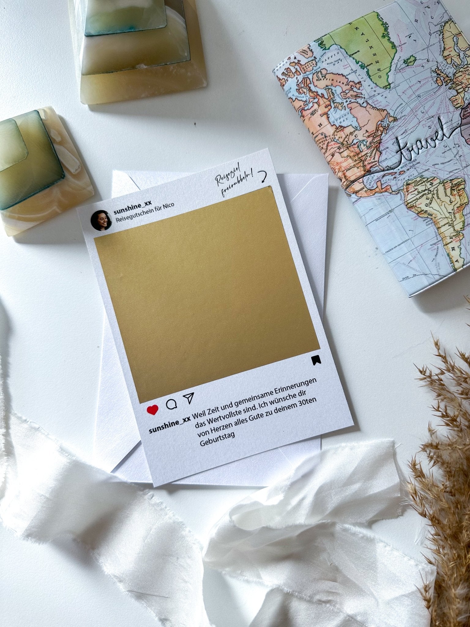 Ein Flatlay-Bild zeigt den „Reisegutschein im Instagram Stil mit Rubbelaufkleber und Umschlag“ von Funkenschauer, mit einer Postkarte mit goldenem Rubbelfeld, einem Rand im Instagram-Stil und deutschem Text. Die Karte liegt auf einem weißen Umschlag und ist von einer kleinen Globuskarte, einem Band und dekorativen Steinen umgeben. Getrocknete Gräser zieren die untere rechte Ecke. Perfekt als personalisiertes Geschenk oder Reisegutschein.