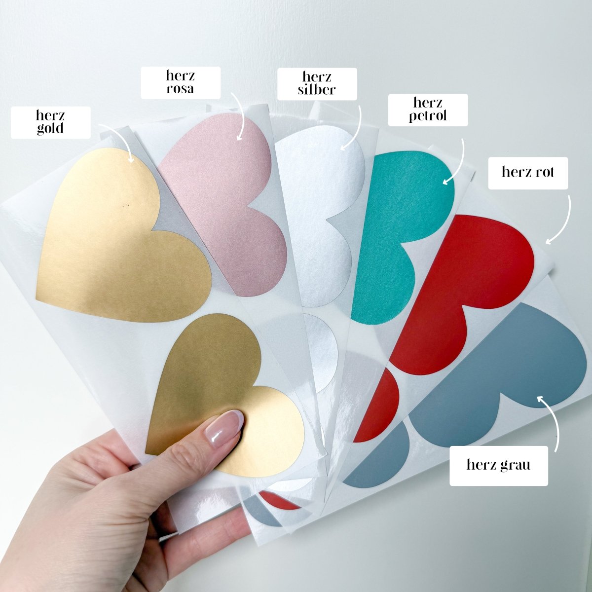 Eine Hand hält große Herzaufkleber in Gold, Rosa, Silber, Türkis, Rot und Grau. Jeder Bogen ist mit dem Farbnamen beschriftet. Auf weißem Hintergrund bieten diese Aufkleber eine personalisierte Geschenkidee für jeden Anlass aus der Kollektion „Reisegutschein zum Verschenken“ von funkenschauer.