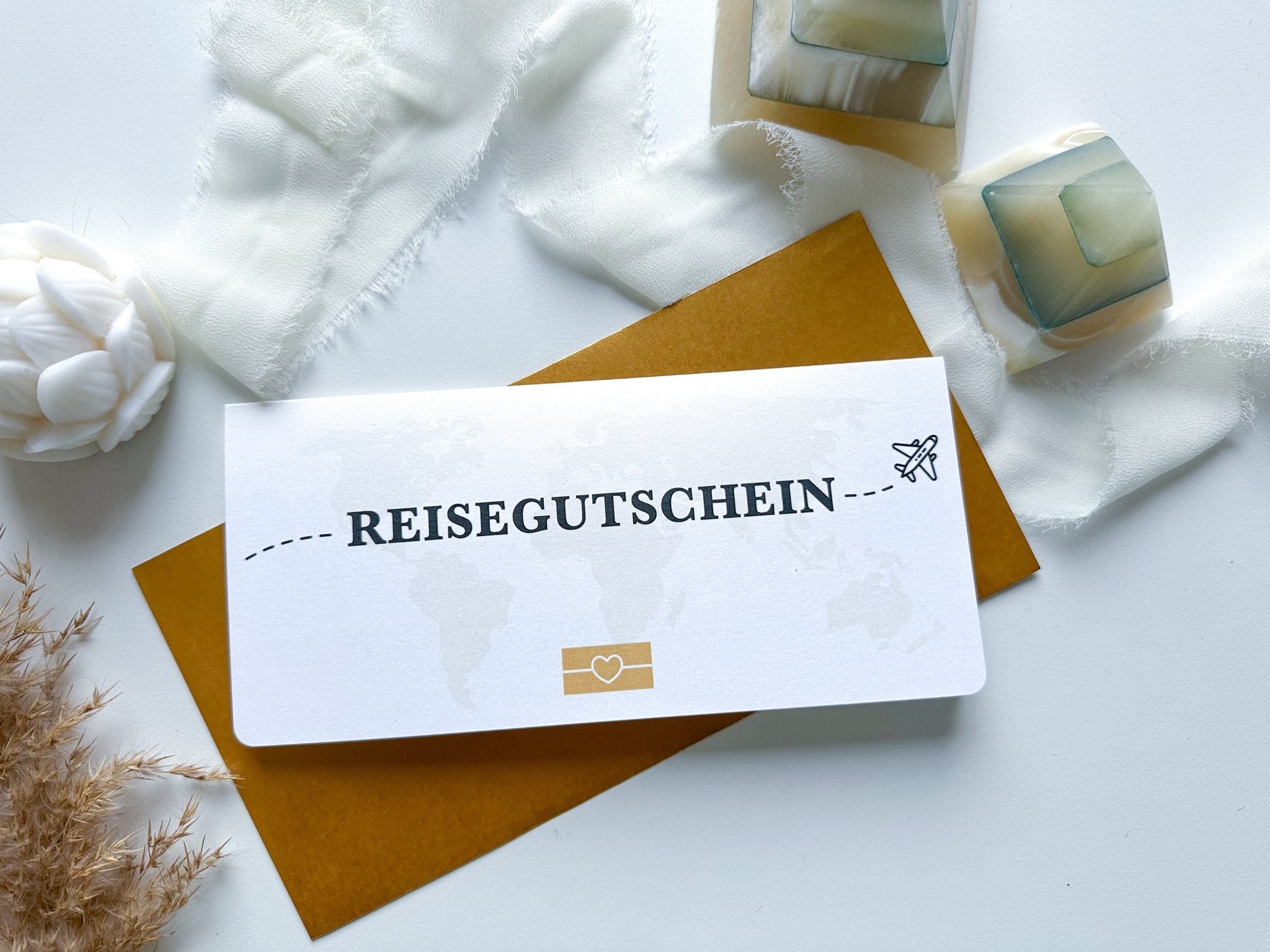 Ein personalisierter Reisegutschein von funkenschauer mit der Aufschrift „Reisegutschein Personalisierbar mit Rubbelfeld zum Aufklappen“ und im Stil eines Flugtickets gestaltet, liegt auf einem braunen Umschlag. Der Hintergrund besteht aus weißem, ausgefranstem Stoff, einem kleinen weißen Blumenornament und zwei Seifenwürfeln, die in durchsichtiges Plastik eingewickelt sind. Die Szene wirkt leicht und luftig.